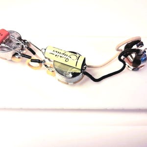 May include: A guitar wiring harness featuring potentiometers, a capacitor, and wiring. The capacitor is orange, and the wiring is black, white, and beige. The components are arranged on a white surface. The image is a close-up.
