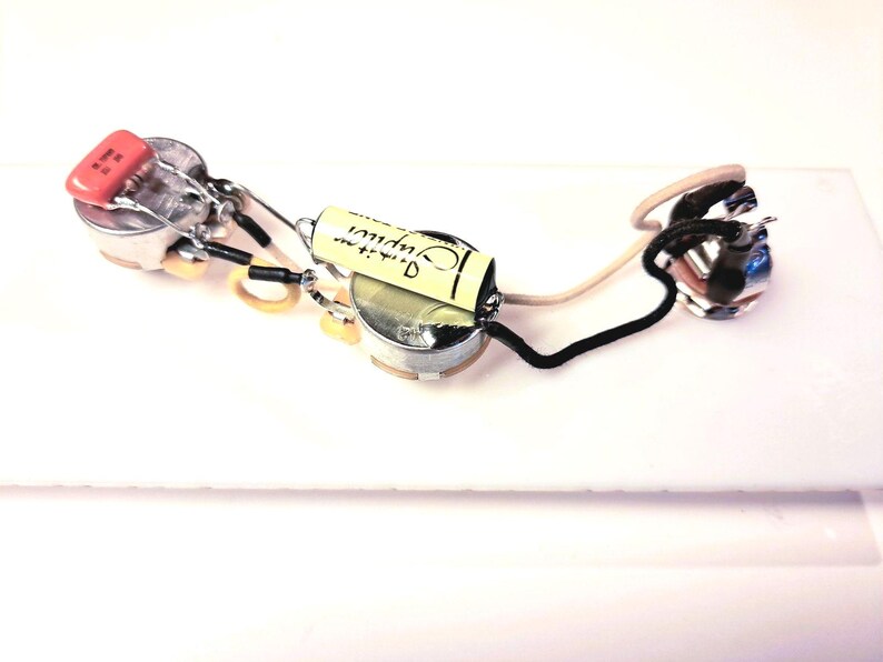 May include: A guitar wiring harness featuring potentiometers, a capacitor, and wiring. The capacitor is orange, and the wiring is black, white, and beige. The components are arranged on a white surface. The image is a close-up.