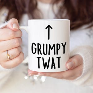 Może przedstawiać: Biały ceramiczny kubek z czarnym napisem "GRUMPY TWAT" ze strzałką skierowaną w górę nad tekstem. Kubek jest trzymany przez osobę ubraną w biały sweter.