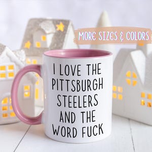 White ceramic mug with a pink interior and handle. The mug features the text "I LOVE THE PITTSBURGH STEELERS AND THE WORD FUCK" in black.