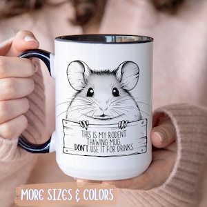 May include: White ceramic mug with a black rim and handle, featuring a black and white illustration of a rodent. The mug has the text "THIS IS MY RODENT THAWING MUG, DON'T USE IT FOR DRINKS". The bottom of the mug has the text "MORE SIZES & COLORS".