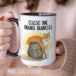 May include: White ceramic mug with a black rim and handle, featuring a cartoon orange tabby cat on a rock. The mug has the text "CLASSIC ONE ORANGE BRAINCELL". The bottom of the mug has the text "MORE SIZES & COLORS".