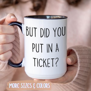 May include: White ceramic mug with black interior and handle, featuring the text "BUT DID YOU PUT IN A TICKET?". The mug is held by a person wearing a pink sweater. The bottom of the mug has the text "MORE SIZES & COLORS".