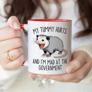 May include: White ceramic mug with a red interior and handle. The mug features a cartoon opossum illustration and the text "MY TUMMY HURTS AND I'M MAD AT THE GOVERNMENT" in black lettering. The mug is held by a person.