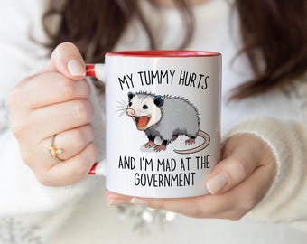 May include: White ceramic mug with a red interior and handle. The mug features a cartoon opossum illustration and the text "MY TUMMY HURTS AND I'M MAD AT THE GOVERNMENT" in black lettering. The mug is held by a person.
