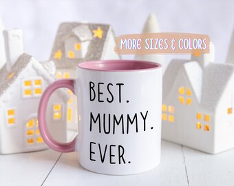 Puede incluir: Taza de cerámica blanca con asa e interior rosa, con el texto "BEST. MUMMY. EVER." en negro. La taza se presenta frente a casas y árboles decorativos blancos.
