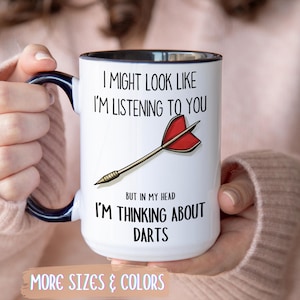 Könnte beinhalten: Weißer Keramikbecher mit schwarzem Innenraum und Henkel, mit dem Text "I MIGHT LOOK LIKE I'M LISTENING TO YOU" und "BUT IN MY HEAD I'M THINKING ABOUT DARTS". Eine Cartoon-Dart-Grafik ist ebenfalls auf dem Becher. Die Worte "MORE SIZES & COLORS" stehen unten.