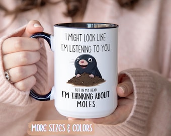 May include: White ceramic mug with a black interior and handle, featuring a cartoon mole illustration. Text reads "I might look like I'm listening to you, but in my head I'm thinking about moles." Additional text at the bottom says "More sizes & colors."