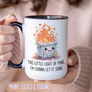 Op de afbeelding: Witte keramische mok met een zwarte handgreep en rand, met een cartoon vuilnisbak in brand met een lachend gezicht. De tekst luidt: "THIS LITTLE LIGHT OF MINE, I'M GONNA LET IT SHINE".