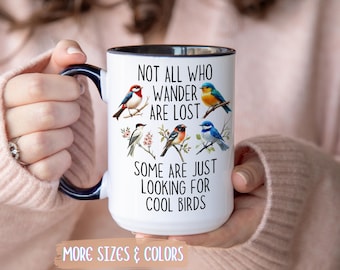 May include: White ceramic mug with a black interior and handle, featuring colorful illustrations of birds and the text "NOT ALL WHO WANDER ARE LOST, SOME ARE JUST LOOKING FOR COOL BIRDS." The mug is held by a person.