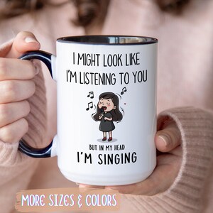 Op de afbeelding: Witte keramische mok met een zwarte binnenkant en handvat, met de tekst "I MIGHT LOOK LIKE I'M LISTENING TO YOU BUT IN MY HEAD I'M SINGING." Een cartoon illustratie van een zingend meisje staat ook op de mok. De woorden "MORE SIZES & COLORS" staan onderaan.