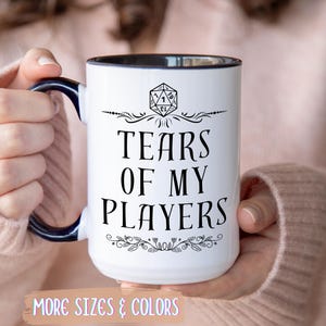 Könnte beinhalten: Weißer Keramikbecher mit schwarzem Rand und Henkel, mit dem Text "TEARS OF MY PLAYERS" in einer kräftigen, stilisierten Schriftart. Der Becher hat ein dekoratives Design mit einem D20-Würfel und floralen Akzenten.