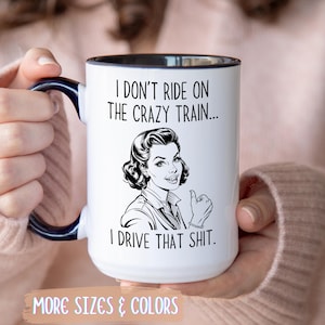 May include: White ceramic mug with a black rim and handle, featuring a retro illustration of a woman giving a thumbs up. The mug has the text "I DON'T RIDE ON THE CRAZY TRAIN... I DRIVE THAT SHIT." The words "MORE SIZES & COLORS" are at the bottom.