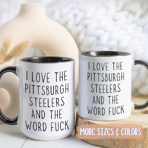 Two white ceramic mugs with black handles and rims. Each mug has the text "I LOVE THE PITTSBURGH STEELERS AND THE WORD FUCK" printed in black.
