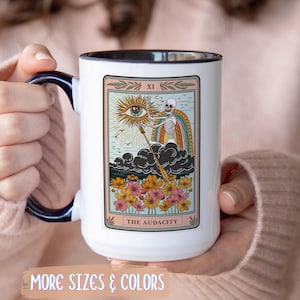 May include: White ceramic mug with a black handle and rim, featuring a tarot card design. The card depicts an eye, skeleton, and rainbow, with the text "THE AUDACITY". The mug is held by a person wearing a pink sweater.