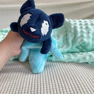 May include: A blue plush toy with a white face and red stitched mouth. The toy is lying on a light blue blanket with a white background.
