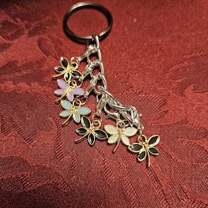 Puede incluir: Un llavero plateado con un anillo plateado y una cadena plateada. Hay seis dijes unidos a la cadena, cada uno con forma de mariposa con esmalte de diferentes colores. Las mariposas son moradas, azules, negras, blancas y doradas.