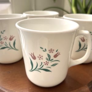 May include: Four off-white ceramic mugs with a floral design featuring pink tulips and green leaves. The mugs have a curved shape with a handle and are arranged on a wooden surface. The mugs are part of a set.