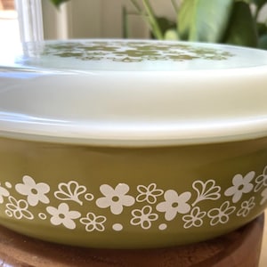 以下が含まれることがあります： ベースと蓋に白い花柄模様が入った、ヴィンテージのオリーブグリーンのキャセロール皿。白い蓋とハンドルが付いています。クラシックなキッチン用品です。