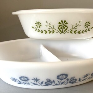 May include: Two white vintage serving dishes. The top dish has a floral design in green. The bottom dish is divided and has a blue floral pattern. Both dishes are oval-shaped and appear to be made of glass.