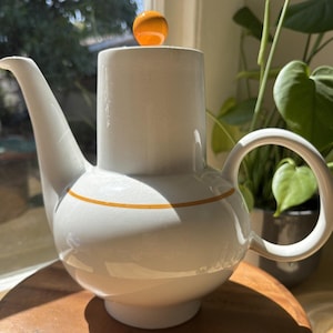 Puede incluir: Tetera de cerámica blanca con base redonda, cuerpo cilíndrico y boquilla curva. Una esfera naranja se encuentra sobre la tapa. La tetera tiene una franja naranja alrededor del centro y un asa redondeada.