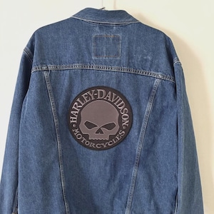 May include: Blue denim jacket with a Harley-Davidson patch featuring a skull and the words "Harley-Davidson Motorcycles" in a circular design. The jacket is hanging on a wooden rack.