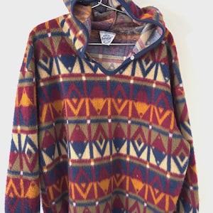 Könnte beinhalten: Ein Vintage-Fleece-Pullover mit einem geometrischen Muster in den Farbtönen Burgunderrot, Marineblau, Gold und Beige. Der Kapuzenpullover hat einen V-Ausschnitt mit Knopfverschluss und marineblauen Besatz. Das Fleece hängt an einem Holzgestell.