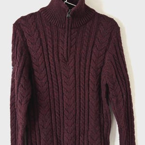 May include: A deep burgundy cable-knit jumper with a high, zipped collar. The jumper is hanging on a white hanger against a white background. The knit pattern features vertical and diagonal textures.