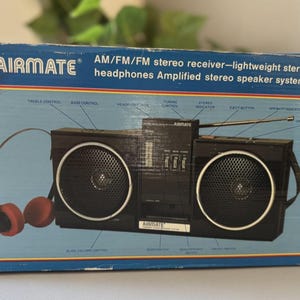 以下が含まれることがあります： オリジナルの箱に入った、ヴィンテージのAIRMATE AM/FMステレオレシーバー。黒いラジオには、2つの大きなスピーカー、ハンドル、アンテナが付いています。赤いヘッドフォンが含まれています。箱は青色で、白とオレンジの文字が書かれています。