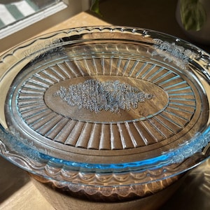May include: Oval glass serving dish with a clear lid. The dish has a light blue tint and decorative handles. The lid features an embossed floral design and radial lines. A wooden surface is visible beneath the glass.