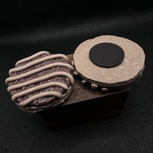May include: Two handmade, decorative magnets resembling cookies. One is a dark brown color with light brown icing stripes. The other is a light brown color with a black magnet on top. Both are on a small, dark wooden block.