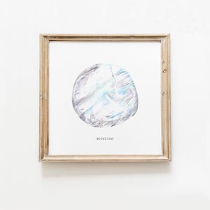 Puede incluir: Una impresión enmarcada de una pintura de acuarela de una piedra lunar. La piedra lunar es de color azul claro y gris con un efecto brillante. La impresión está enmarcada en un marco de madera clara y tiene la palabra "MOONSTONE" impresa debajo de la imagen.