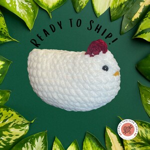 May include: A handmade white crochet chicken with a red comb and a yellow beak. The chicken has a black eye and is surrounded by green leaves on a dark green background. Text on the image reads "READY TO SHIP!"