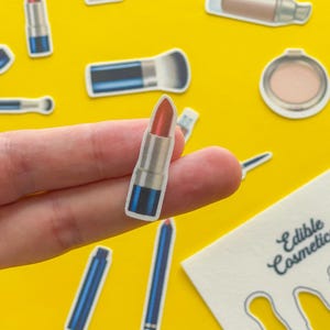 May include: A collection of makeup stickers, including lipstick, mascara, and blush, are scattered on a yellow background. A lipstick sticker is held by a hand. The text "Edible Cosmetics" is visible on a white card.