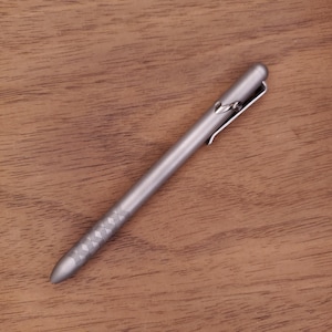 May include: A silver metal pen with a pocket clip and a decorative diamond pattern near the tip. The pen is lying on a wooden surface.