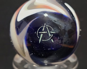 May include: A white, blue, and orange swirl glass marble with a silver anarchy symbol etched on the surface.