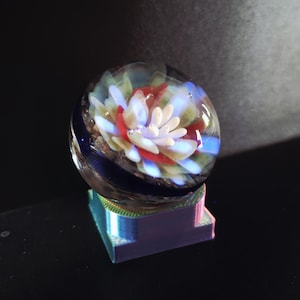 May include: A clear glass orb with a colorful floral design inside, resting on a square, iridescent base. The flower features white, red, and blue petals. The base has a rainbow sheen, reflecting various colors. The background is dark.