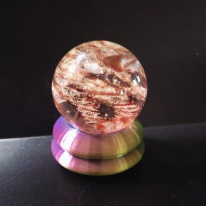 May include: A clear, spherical paperweight with red and gold inclusions, resting on a metallic, rainbow-colored base. The orb appears to have a swirling, textured interior. The base has a layered, circular design.