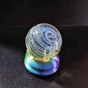 May include: A clear glass sphere with a blue and white spiral design, resting on a rainbow-colored, layered base. The sphere appears to have a galaxy-like pattern inside. The base is composed of concentric rings of color.
