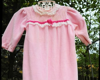 Puede incluir: Un pelele de bebé de terciopelo rosa con ribete de encaje blanco y un lazo rosa. El pelele tiene un cierre de presión en la entrepierna.
