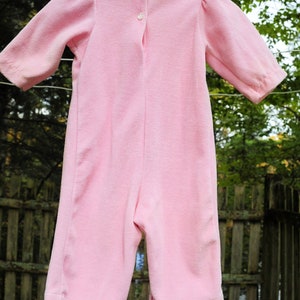 Puede incluir: Un pelele rosa para beb&eacute; con un ribete de encaje blanco alrededor del escote. El pelele tiene un cierre de bot&oacute;n en la espalda.