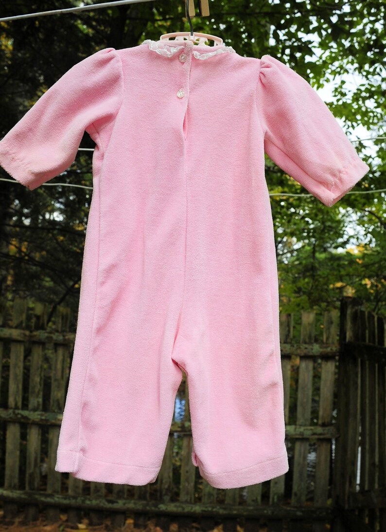 Puede incluir: Un pelele rosa para beb&eacute; con un ribete de encaje blanco alrededor del escote. El pelele tiene un cierre de bot&oacute;n en la espalda.