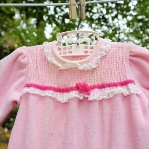 Puede incluir: Un pelele rosa para beb&eacute; con ribete de encaje blanco y un lazo de cinta rosa. El pelele est&aacute; colgado de una percha.