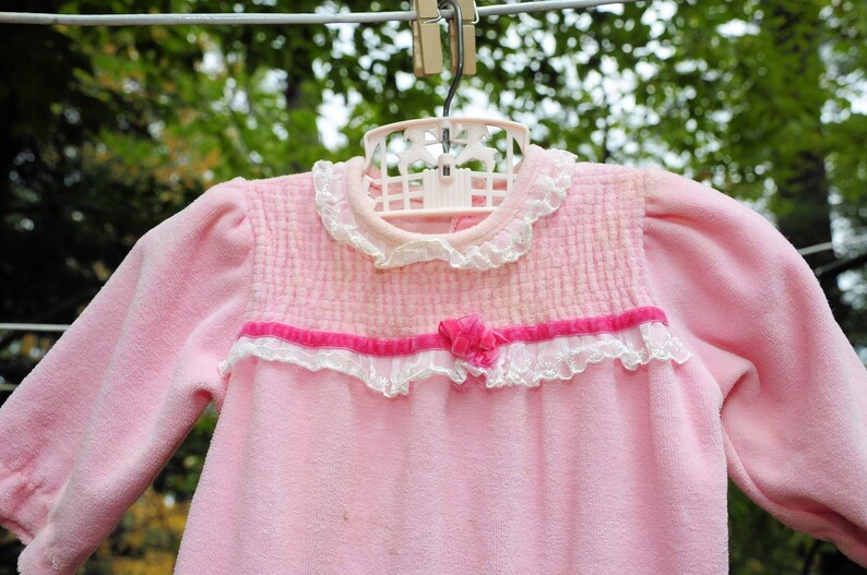 Puede incluir: Un pelele rosa para beb&eacute; con ribete de encaje blanco y un lazo de cinta rosa. El pelele est&aacute; colgado de una percha.