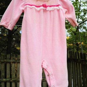 Puede incluir: Un pelele de beb&eacute; de terciopelo rosa con ribete de encaje blanco y un lazo rosa. El pelele tiene cierre de presi&oacute;n en la entrepierna y botones en la parte delantera.