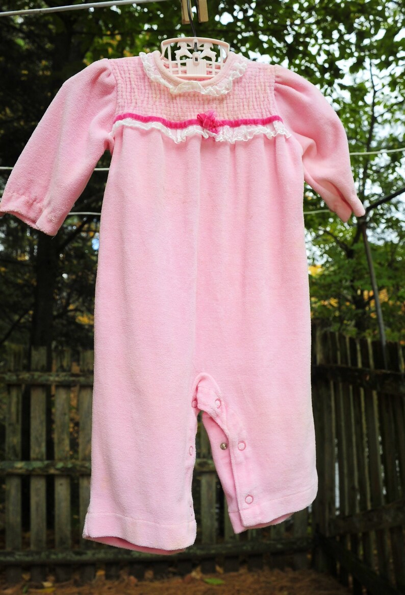 Puede incluir: Un pelele de beb&eacute; de terciopelo rosa con ribete de encaje blanco y un lazo rosa. El pelele tiene cierre de presi&oacute;n en la entrepierna y botones en la parte delantera.