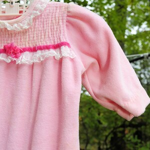 Puede incluir: Una bata de beb&eacute; de terciopelo rosa con ribete de encaje blanco y una cinta rosa. La bata est&aacute; colgada de un tendedero.