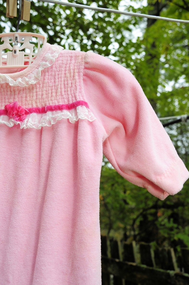 Puede incluir: Una bata de beb&eacute; de terciopelo rosa con ribete de encaje blanco y una cinta rosa. La bata est&aacute; colgada de un tendedero.