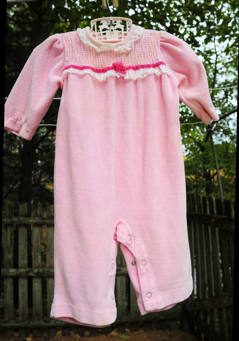 Puede incluir: Un pelele de beb&eacute; de terciopelo rosa con ribete de encaje blanco y un lazo rosa. El pelele tiene un cierre de presi&oacute;n en la entrepierna.
