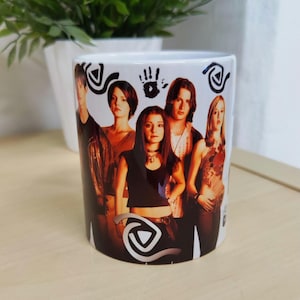 Può includere: Tazza in ceramica bianca con l'immagine di un gruppo di persone. La tazza ha un design avvolgente con un gruppo di persone al centro e simboli decorativi. La tazza è cilindrica e poggia su una superficie chiara.